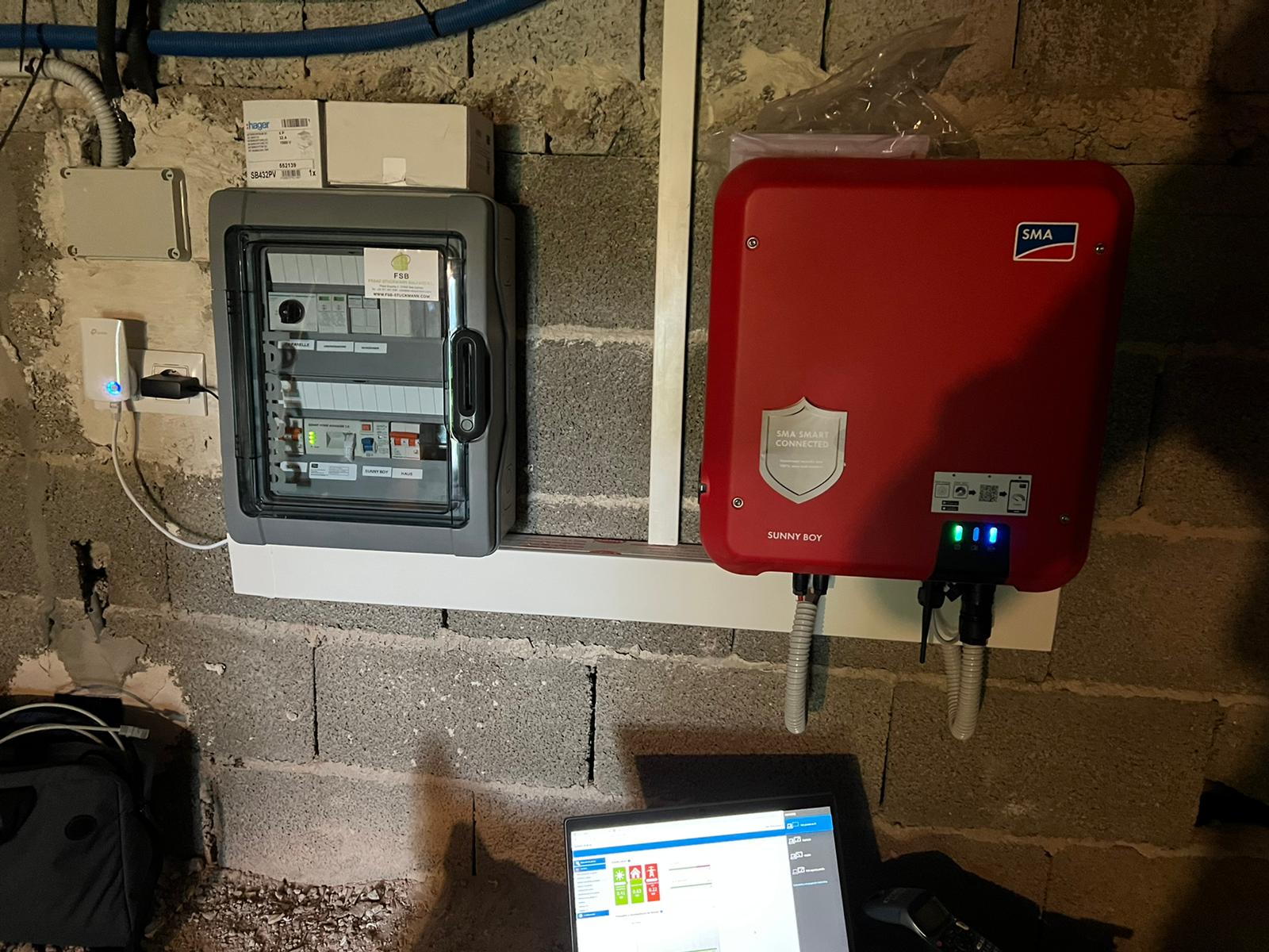 Panel eléctrico e inversor solar rojo montados en una pared de bloques de hormigón. También se ven cables y una computadora portátil.