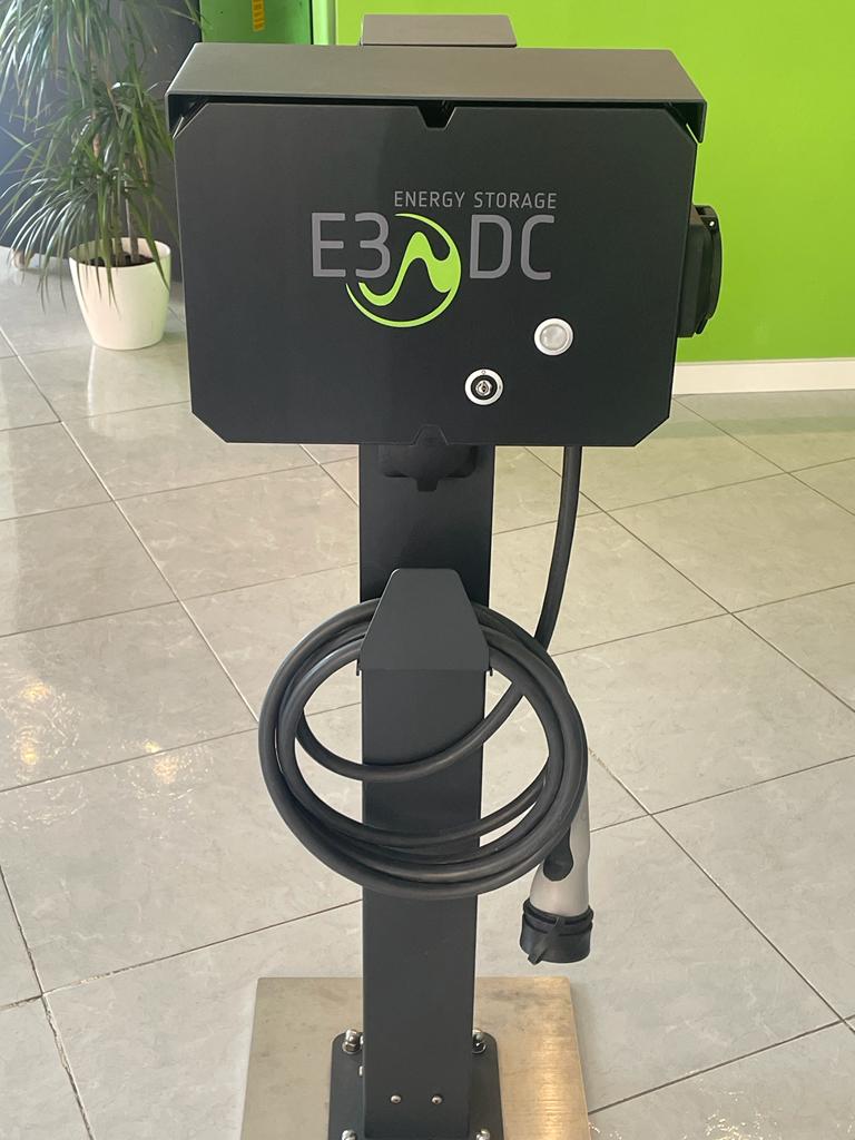 Estación de carga para vehículos eléctricos de color negro, con cable enrollado y logotipo verde, situada en el interior.