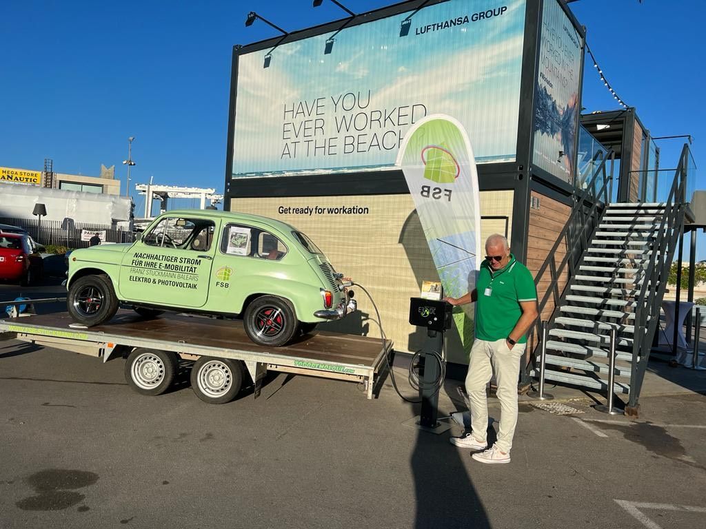 Un automóvil antiguo de color verde claro sobre un remolque con un hombre de pie junto a él, debajo de un gran anuncio, en un día soleado.