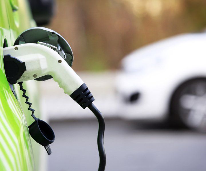 Vehículo eléctrico verde cargándose, con cable de carga blanco conectado. Coche blanco difuminado al fondo.