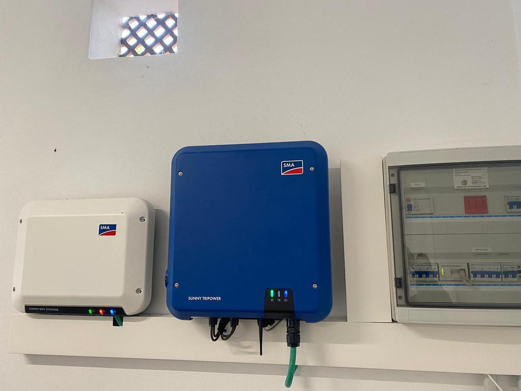 Pared blanca con inversores solares y panel eléctrico. Uno azul, otro blanco y luces indicadoras verdes.