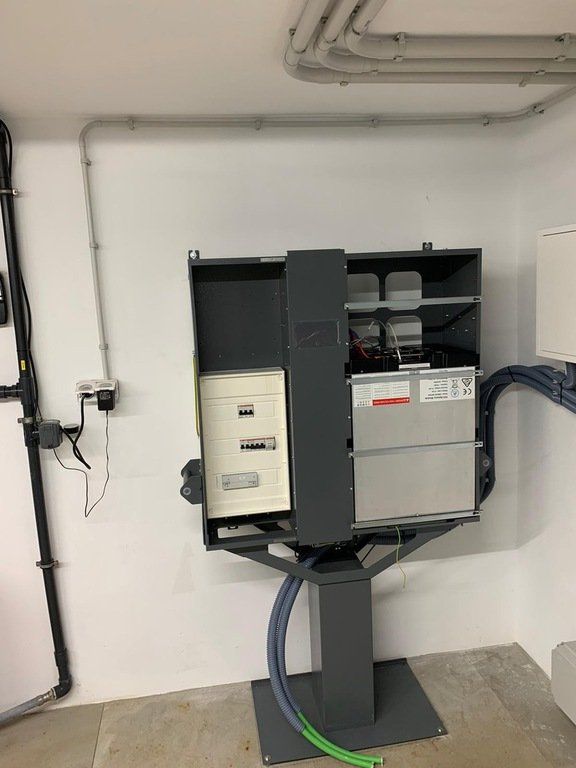 Gabinete eléctrico gris oscuro montado sobre un pedestal en una habitación con paredes, cables y tuberías blancas.