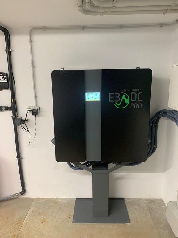 Estación de energía E3/DC Pro negra sobre un pedestal gris, montada en una pared blanca en una habitación, con pantalla y cables conectados.
