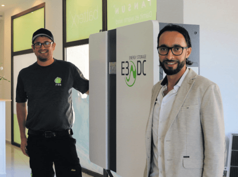 Dos hombres están junto a una unidad de almacenamiento de energía E3/DC blanca. Uno lleva ropa de trabajo; el otro, una chaqueta.