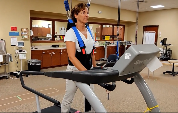 A woman is walking on a treadmill with a harness on her back.