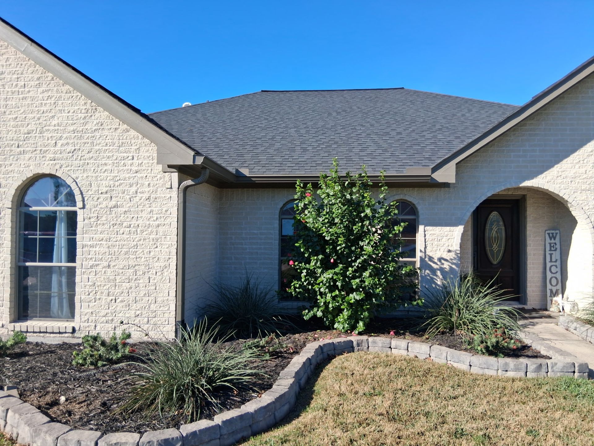 Beige brick house with arched windows, dark roof, front yard landscaping, and blue sky.