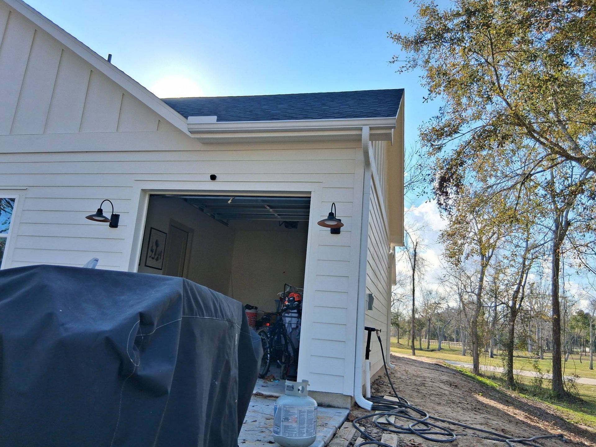 White house exterior with open garage, black grill cover, and trees on a sunny day.