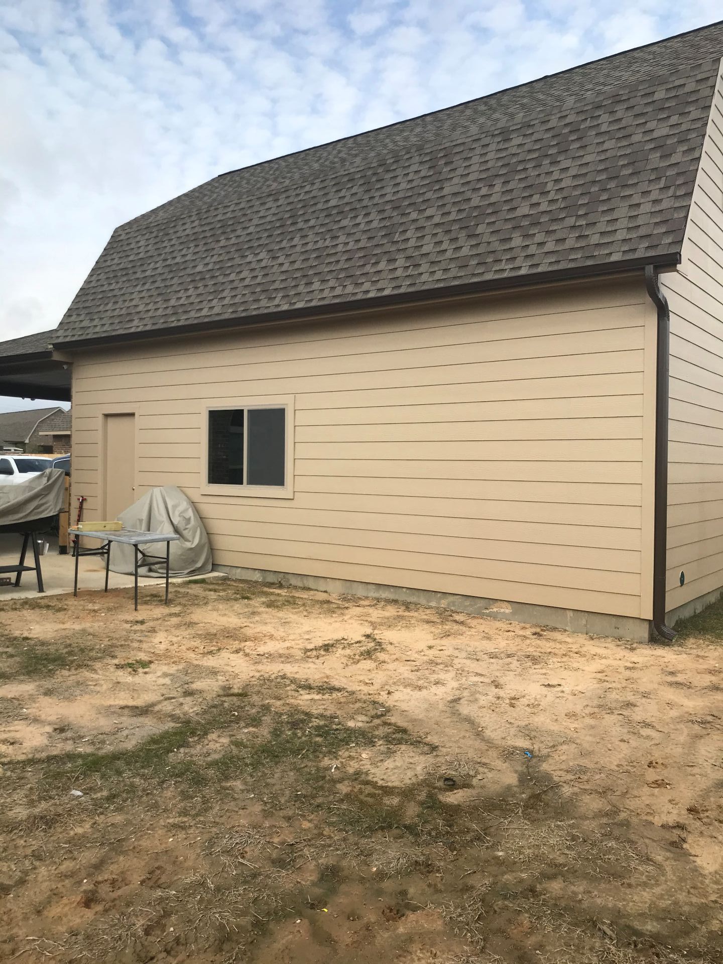 Beige siding building with dark roof, small window and door. Outdoors on dirt ground.