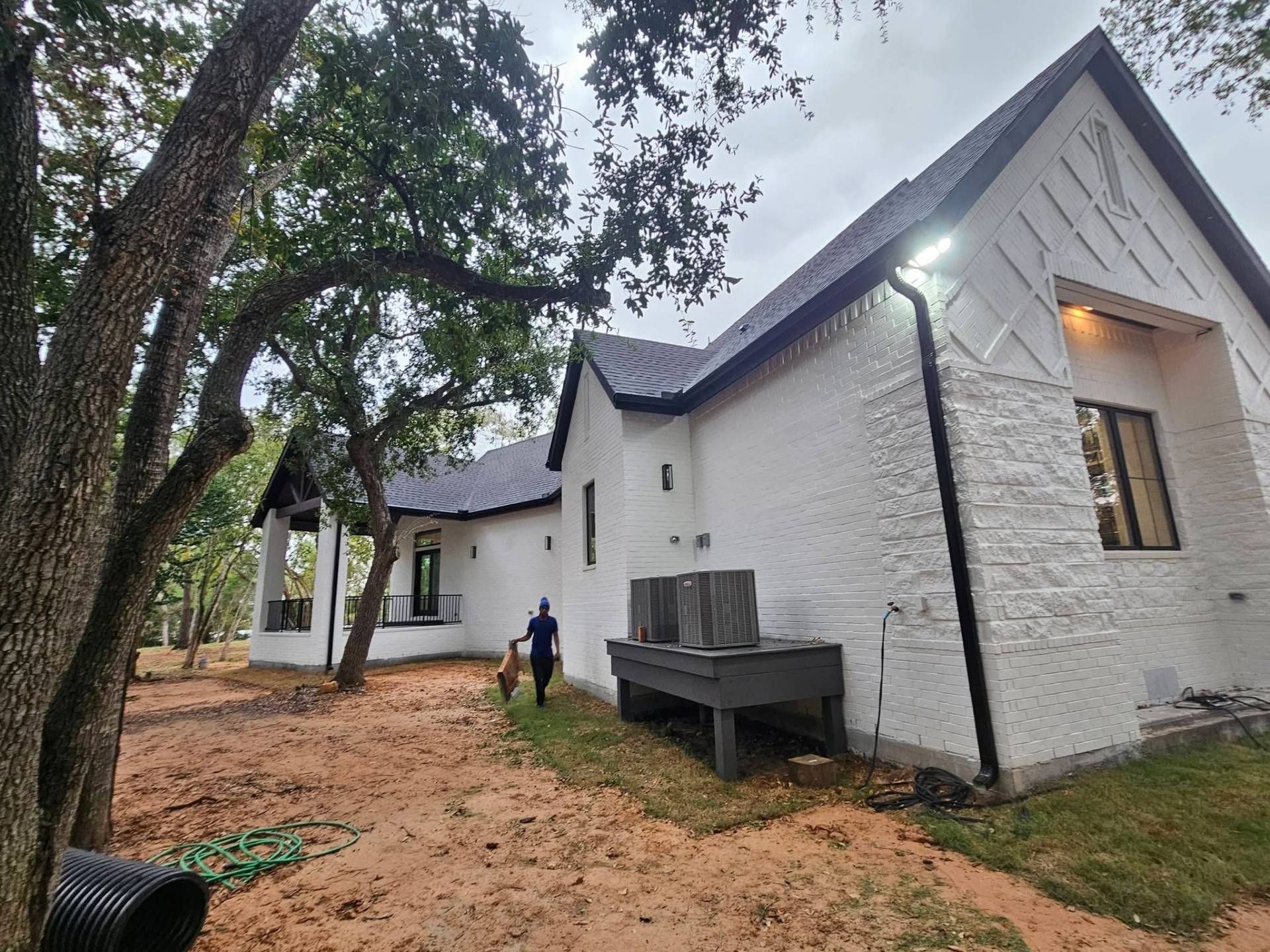 White brick house with black roof, person walking dog, brown dirt, overcast sky.
