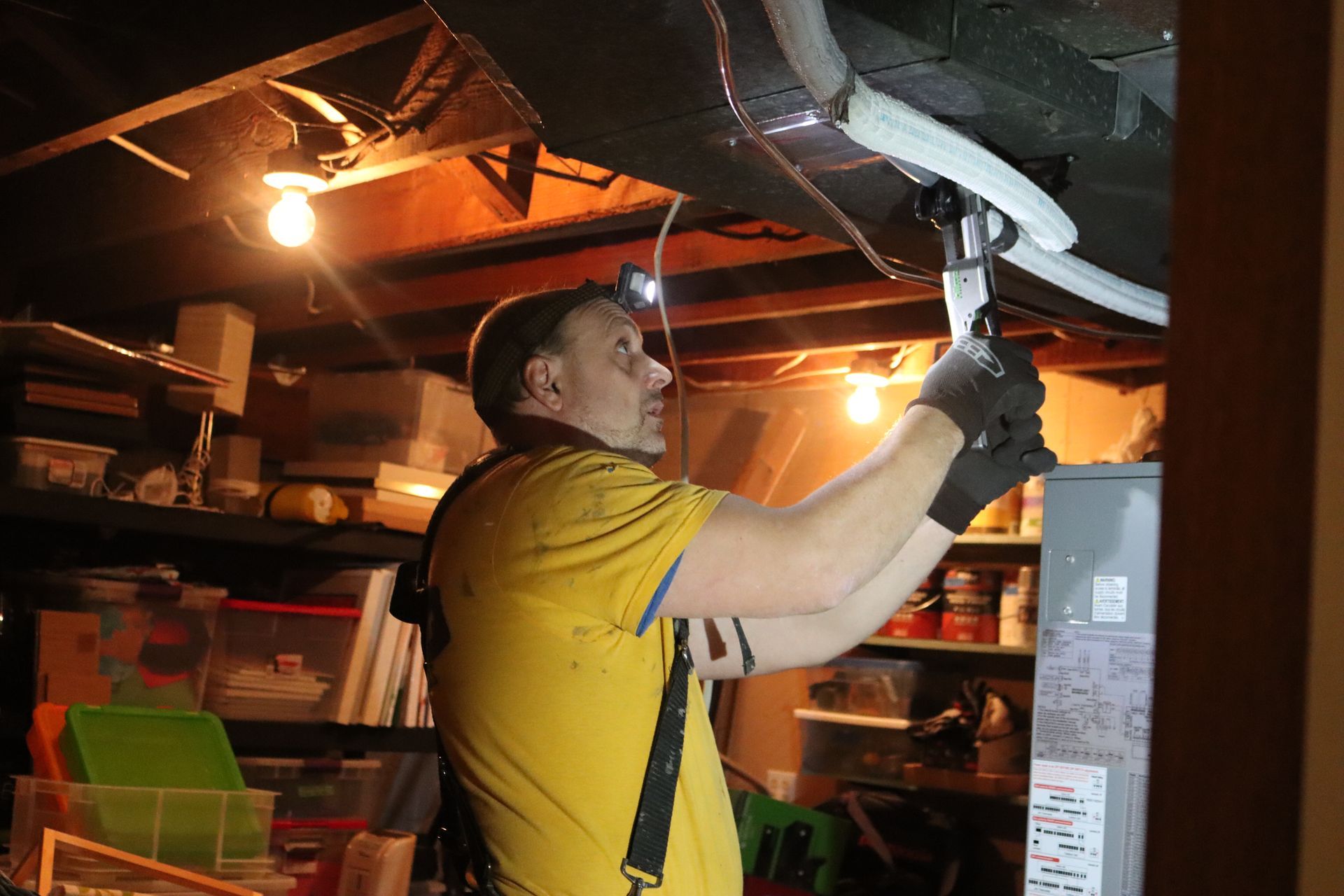Man in yellow shirt, using a tool on equipment in a cluttered basement.