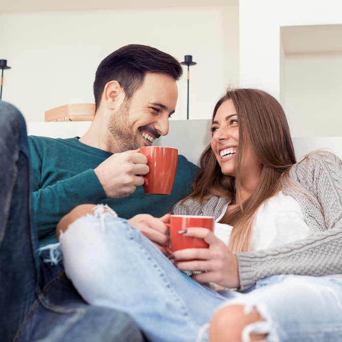 Two people smiling on a couch, holding red mugs in a cozy living room
