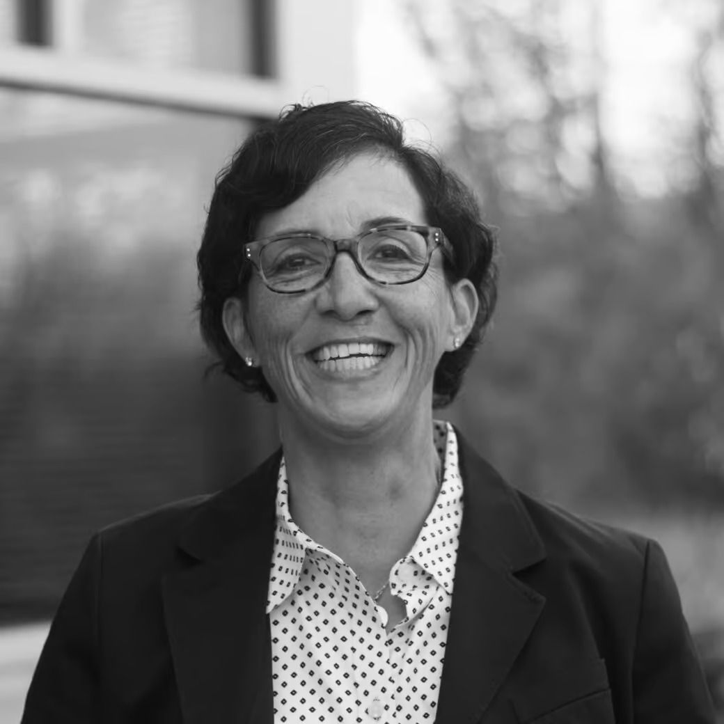 Woman smiling, wearing glasses and a blazer, standing outside a building.