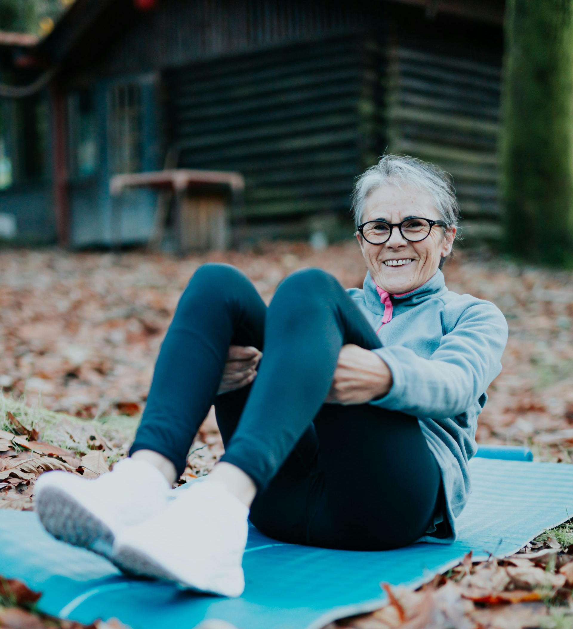 Woman smiling, sitting with knees bent, holding them, on a yoga mat outdoors.