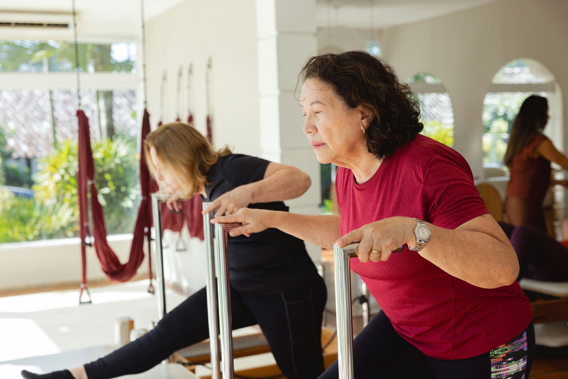 Two women doing exercises, holding onto bars in a studio. One woman in red shirt, one in black. Bright, open space.