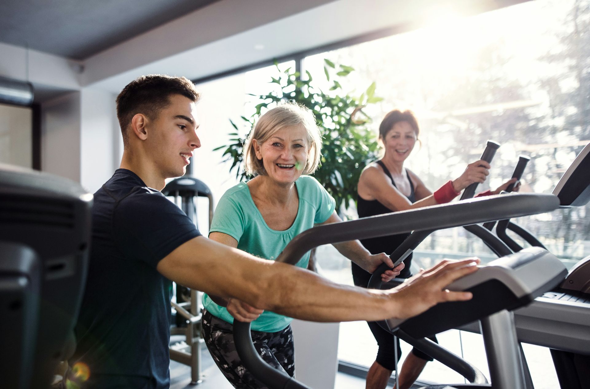Man assisting two women on elliptical trainers in a gym, all smiling. Bright, natural light.
