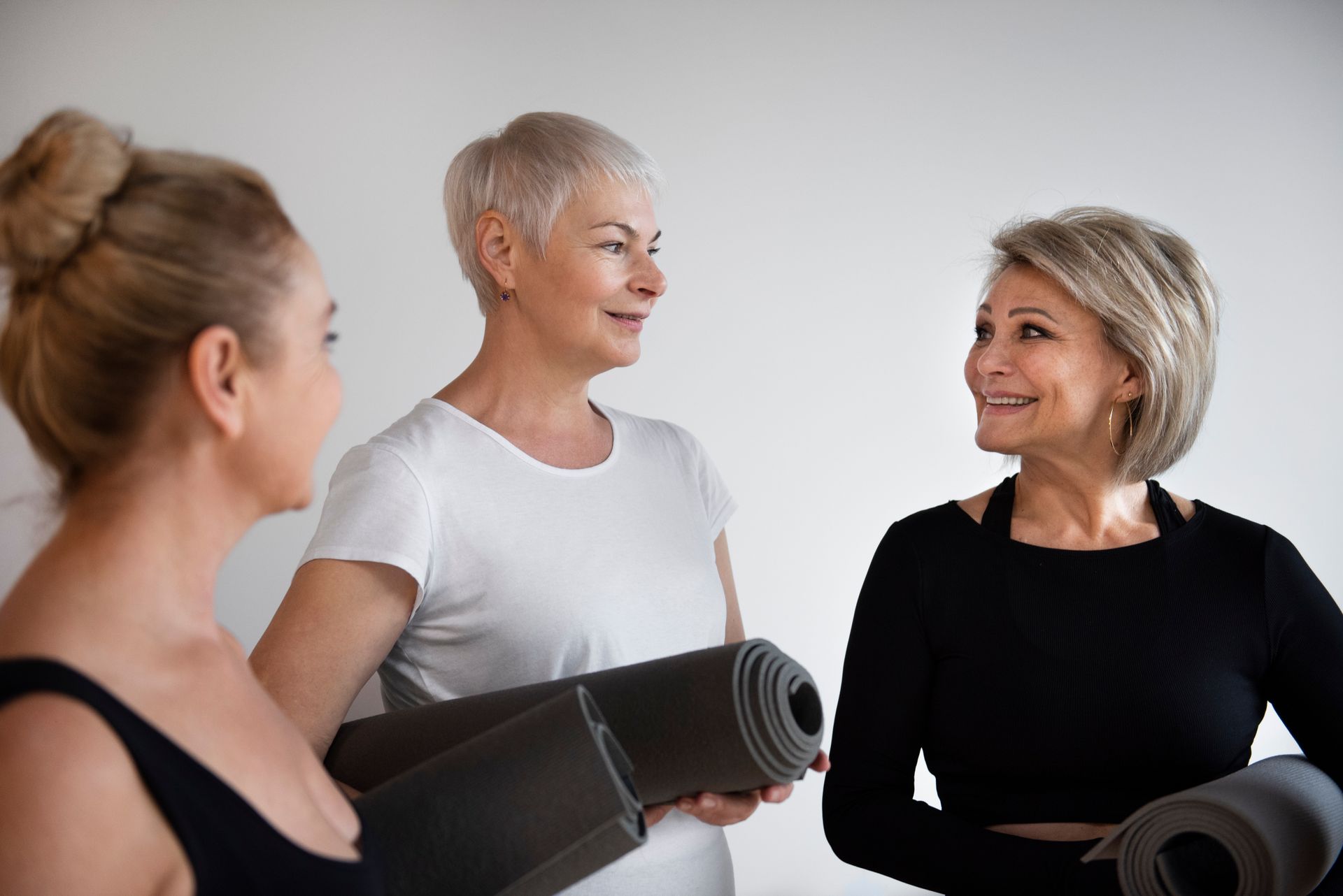 Three women in workout clothes talking, holding rolled yoga mats in a bright room.