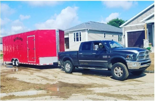 Dark truck towing a red trailer on a gravel driveway, in front of a house on a sunny day.
