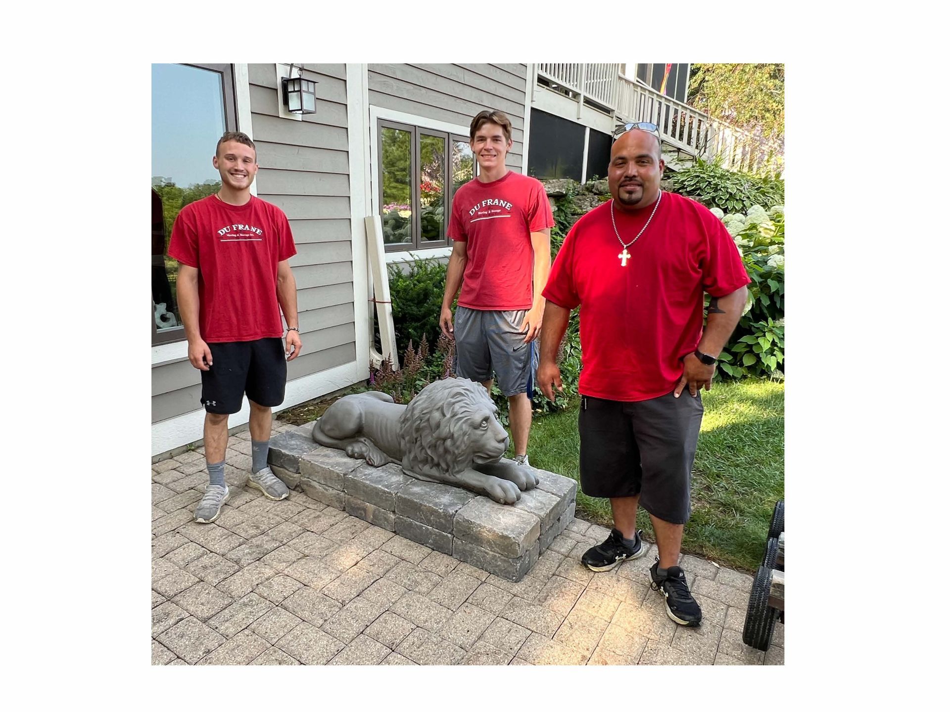Three people stand with a stone lion statue on a patio. All are wearing red shirts.