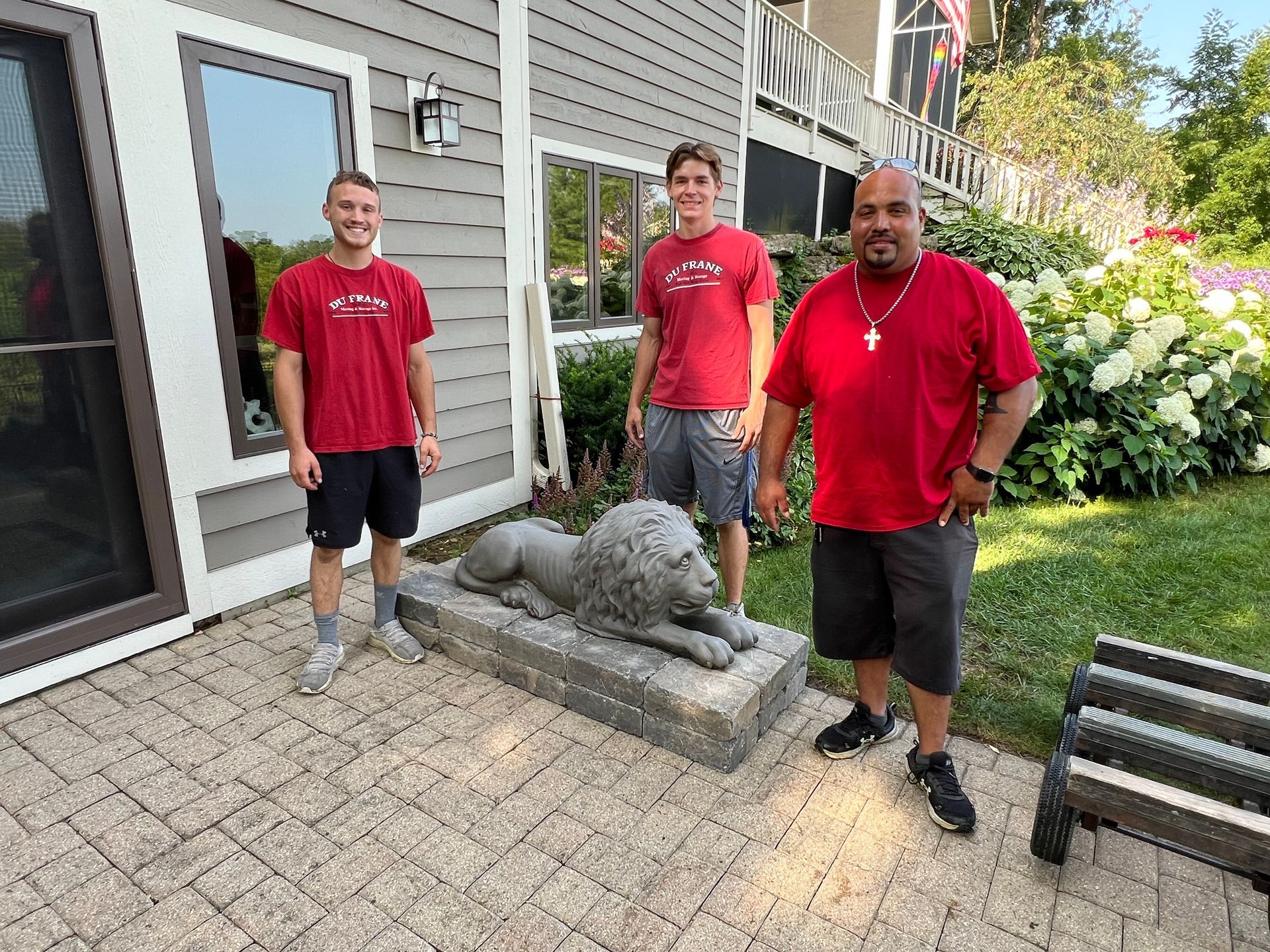 Three people pose next to a stone lion statue on a patio. They wear red shirts and shorts.