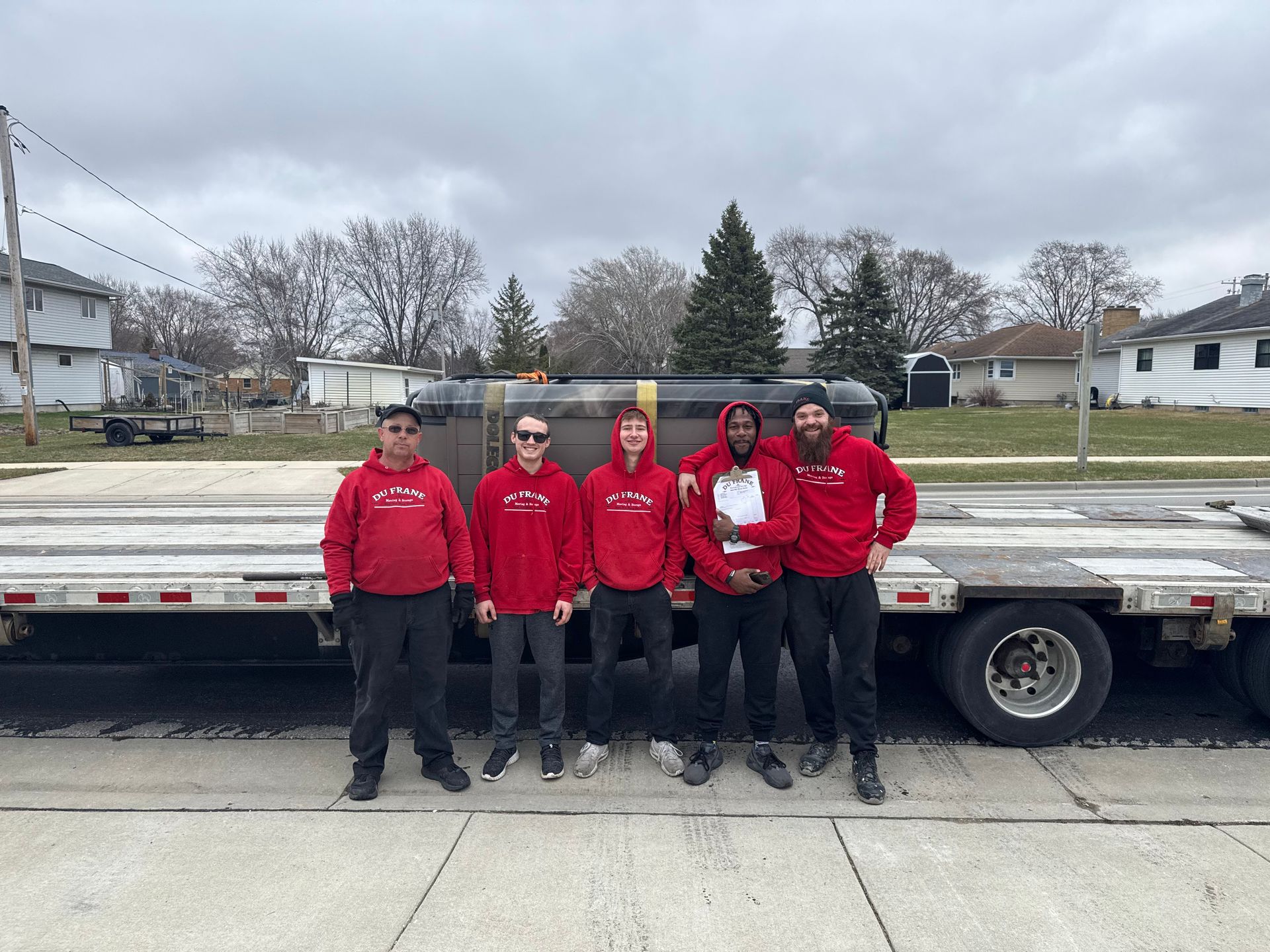 Six people in red hoodies pose next to a flatbed trailer on a street.