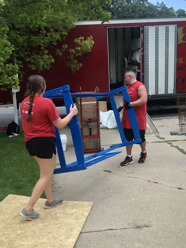 Two people wearing red shirts carry a blue object from a truck onto a wooden platform.