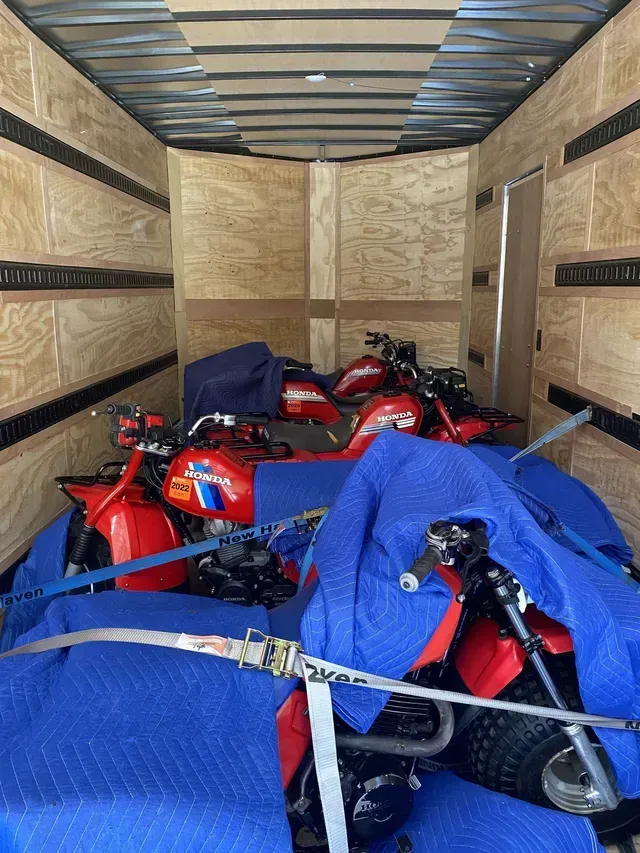 Four red ATVs secured with blue blankets and straps inside a wooden-paneled cargo trailer.