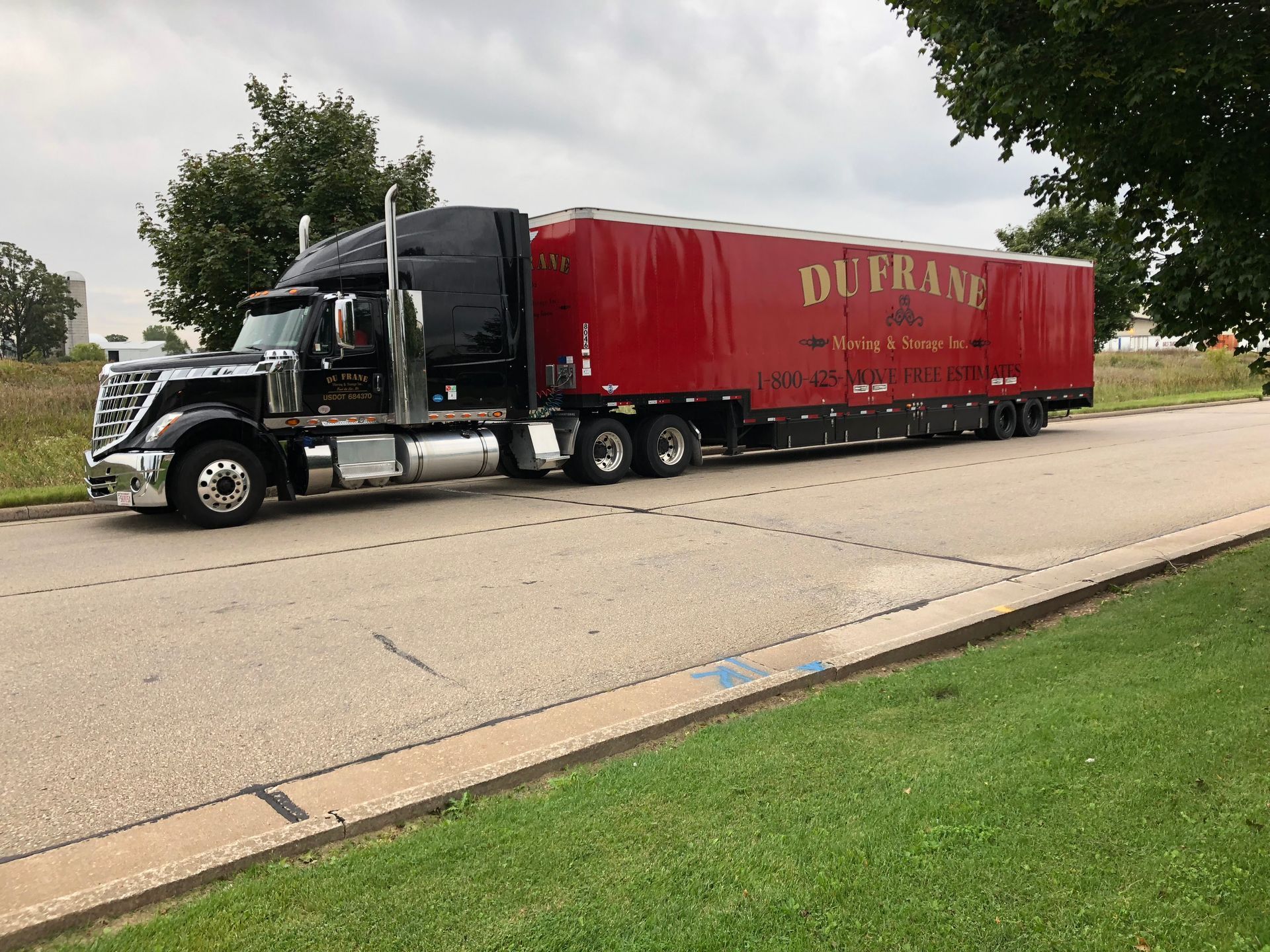 Black semi-truck with red trailer on a road next to a grassy area. The trailer reads