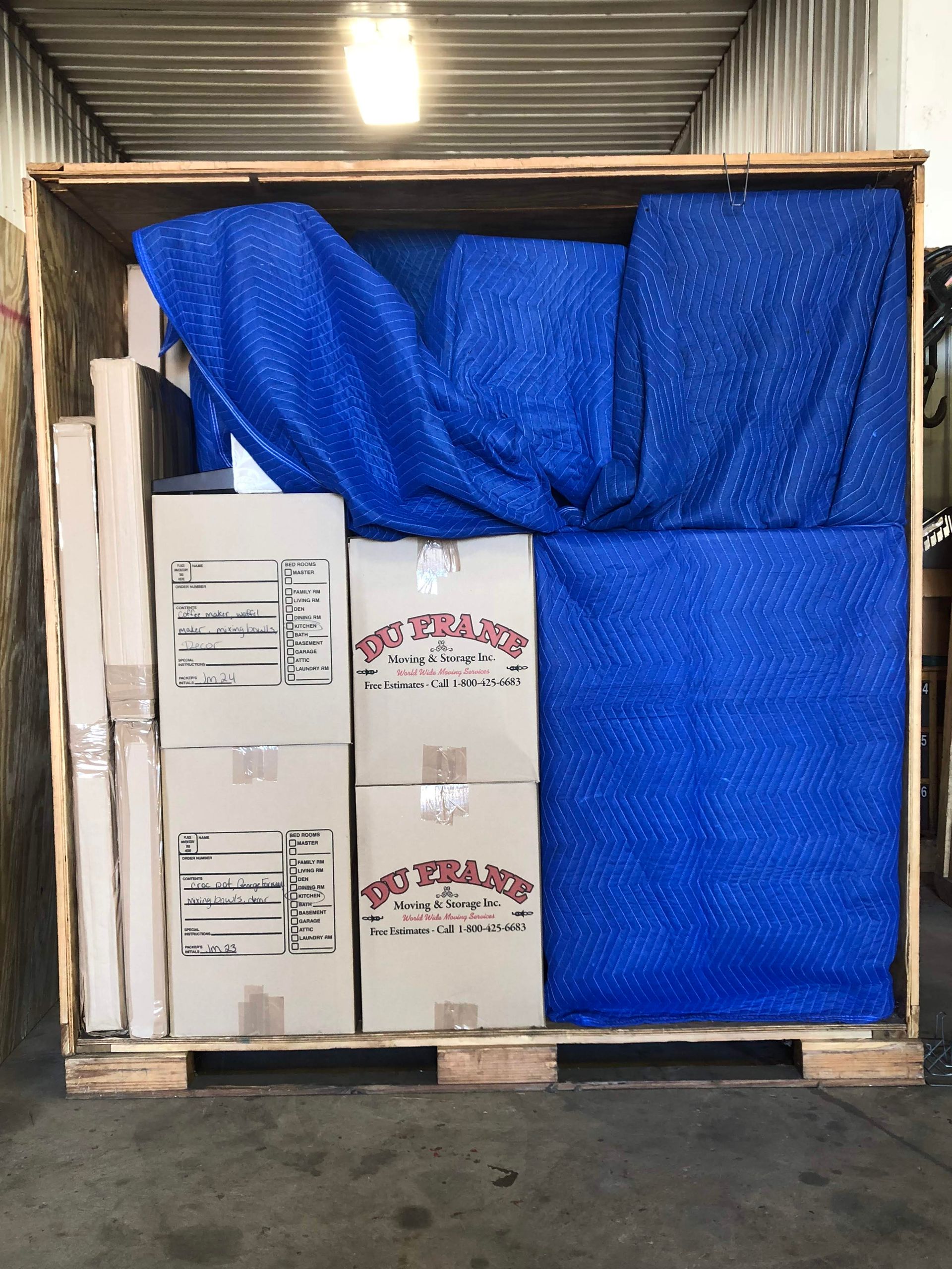 Wooden crate filled with packed boxes and blue protective padding, inside warehouse.