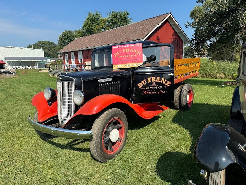 Vintage black-and-red pickup truck on grass beside a red barn at an outdoor event