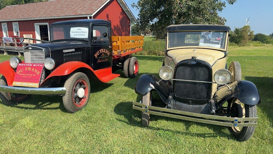 Two vintage pickup trucks on grass, one red and black, one beige and black, in front of a barn.