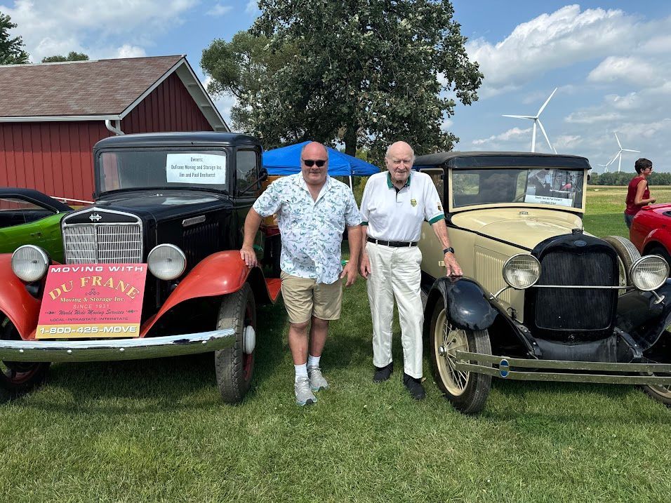 Two men posing between two vintage cars on a grassy field, with a barn and wind turbines behind them.