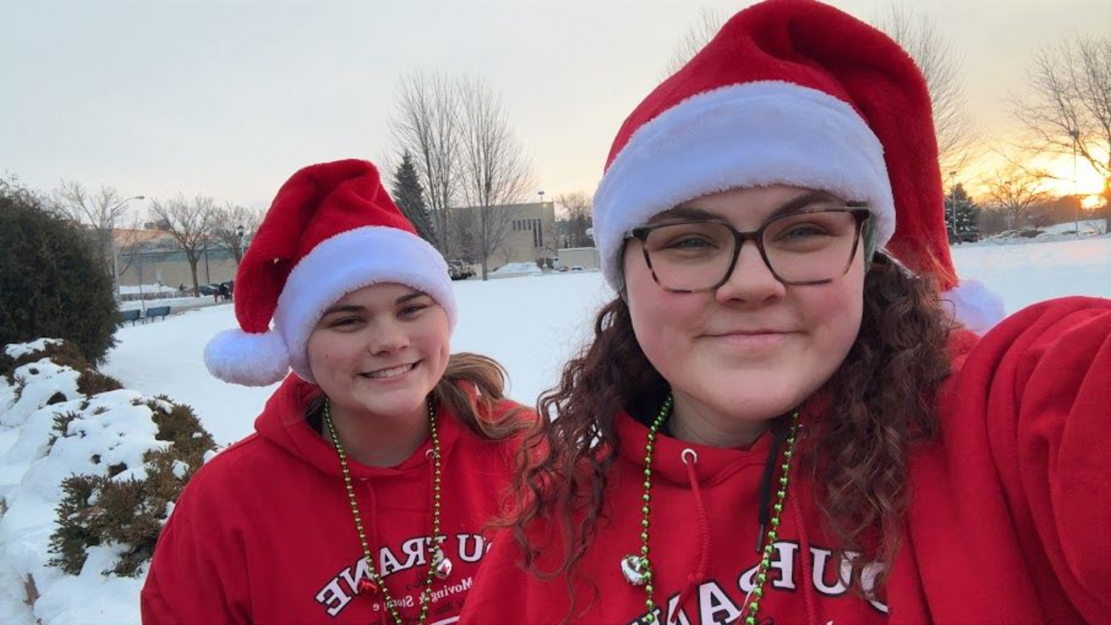 Two people in red Santa hats and red hoodies smiling outdoors in a snowy sunset scene