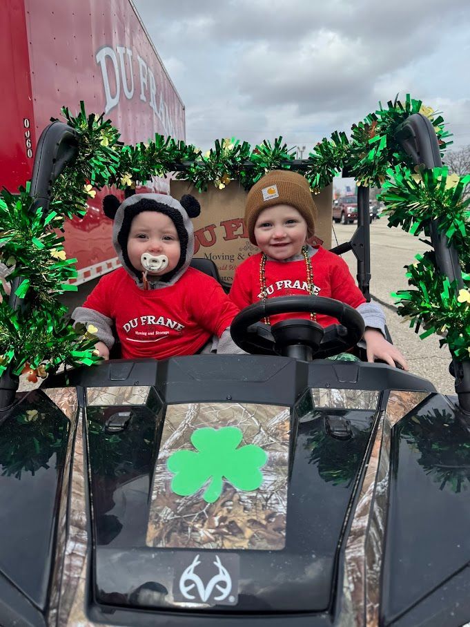 Two children in a black toy car decorated with green shamrocks, smiling at a St. Patrick’s Day event.