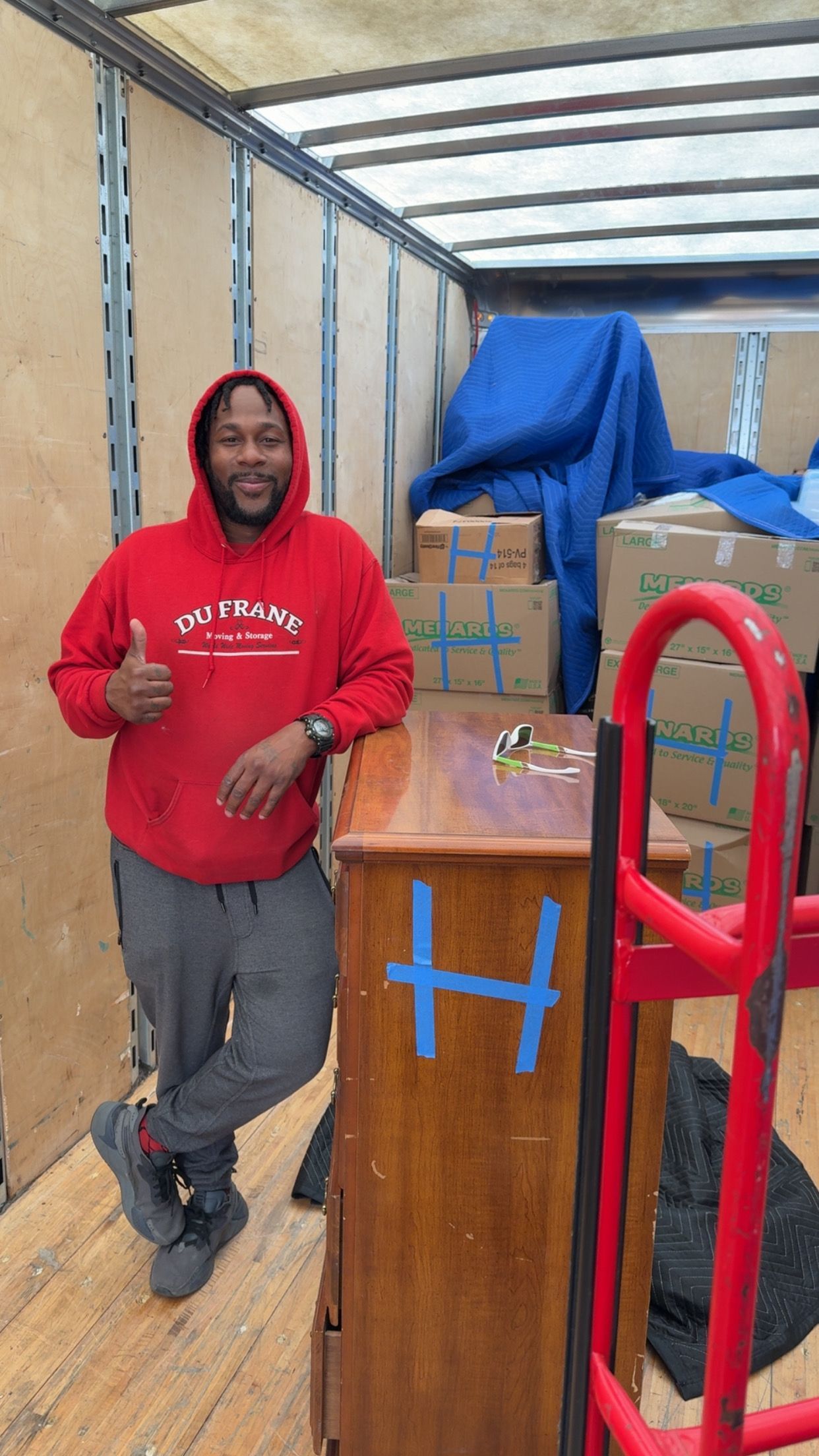 Man in red hoodie inside moving truck giving thumbs up, boxes and furniture visible.