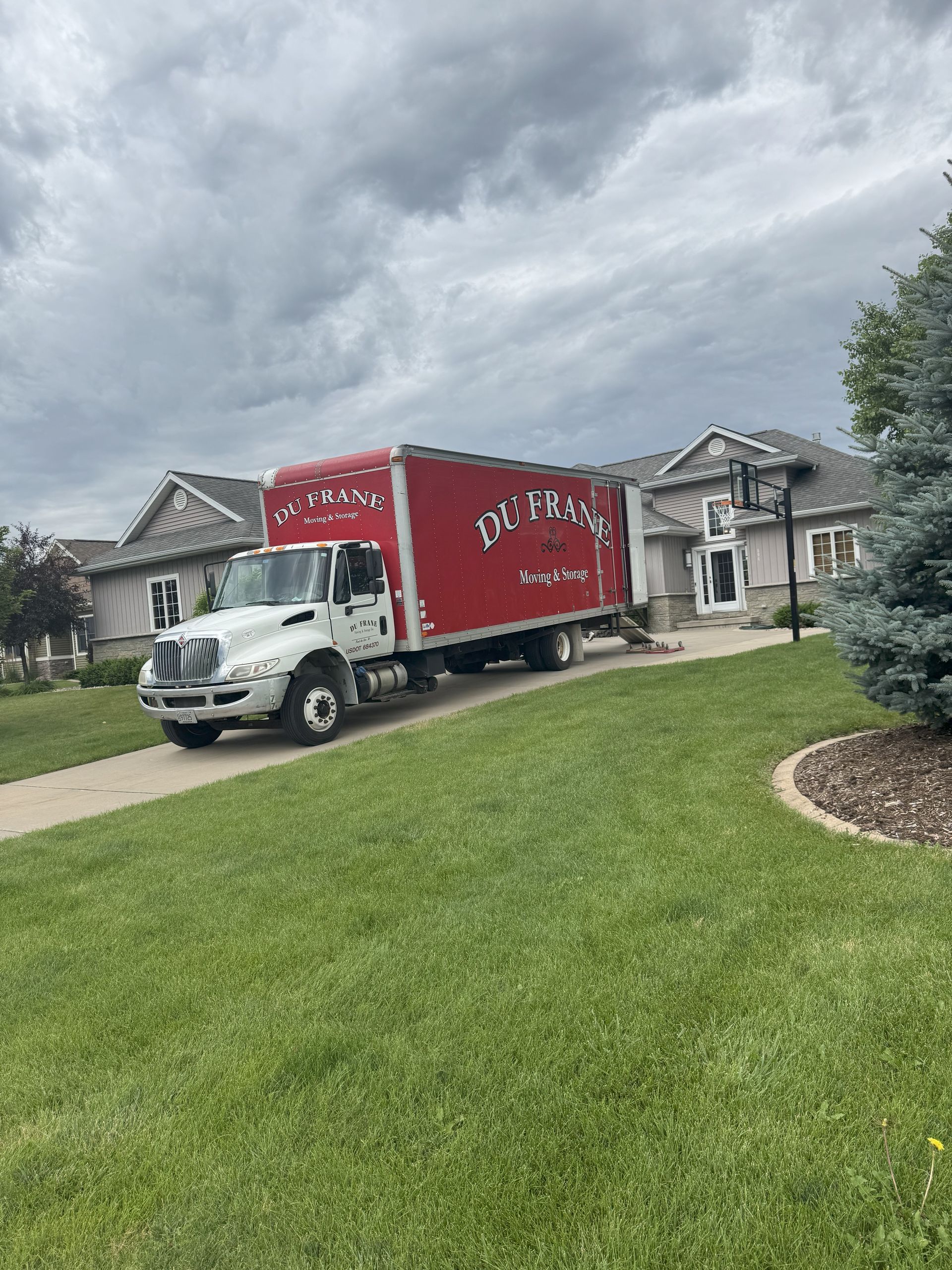 Moving truck in a suburban driveway under cloudy skies, with houses and a green lawn nearby
