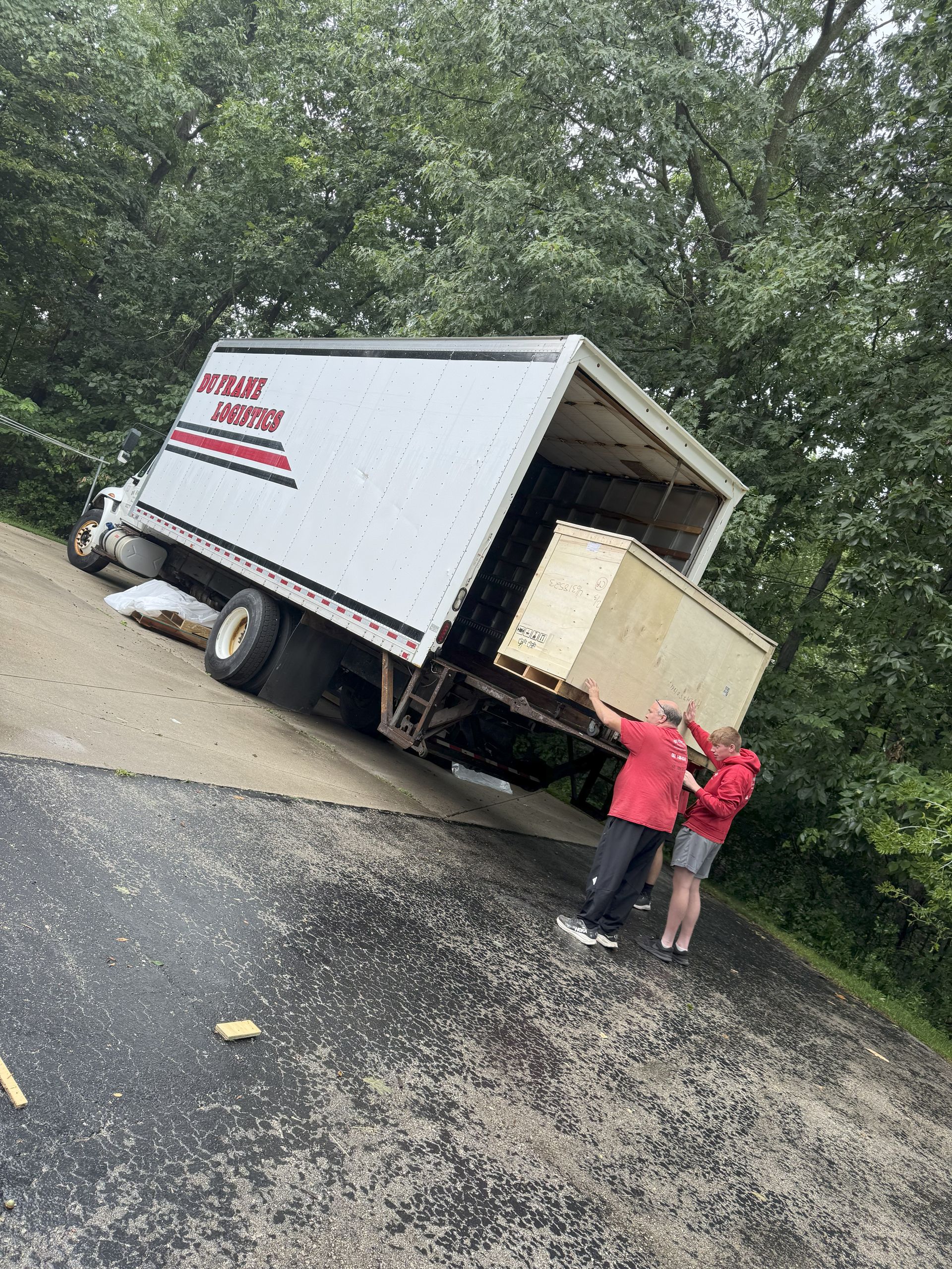 Two workers loading a white moving truck on a roadside by a wooded area