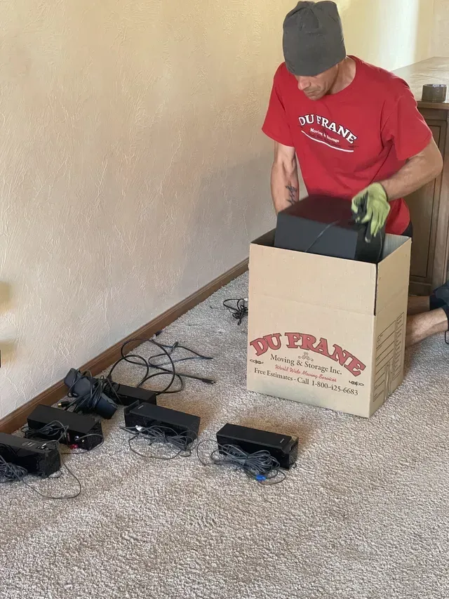 Person packing electronics into a cardboard box on a carpeted floor. Several black devices and cords are on the floor.