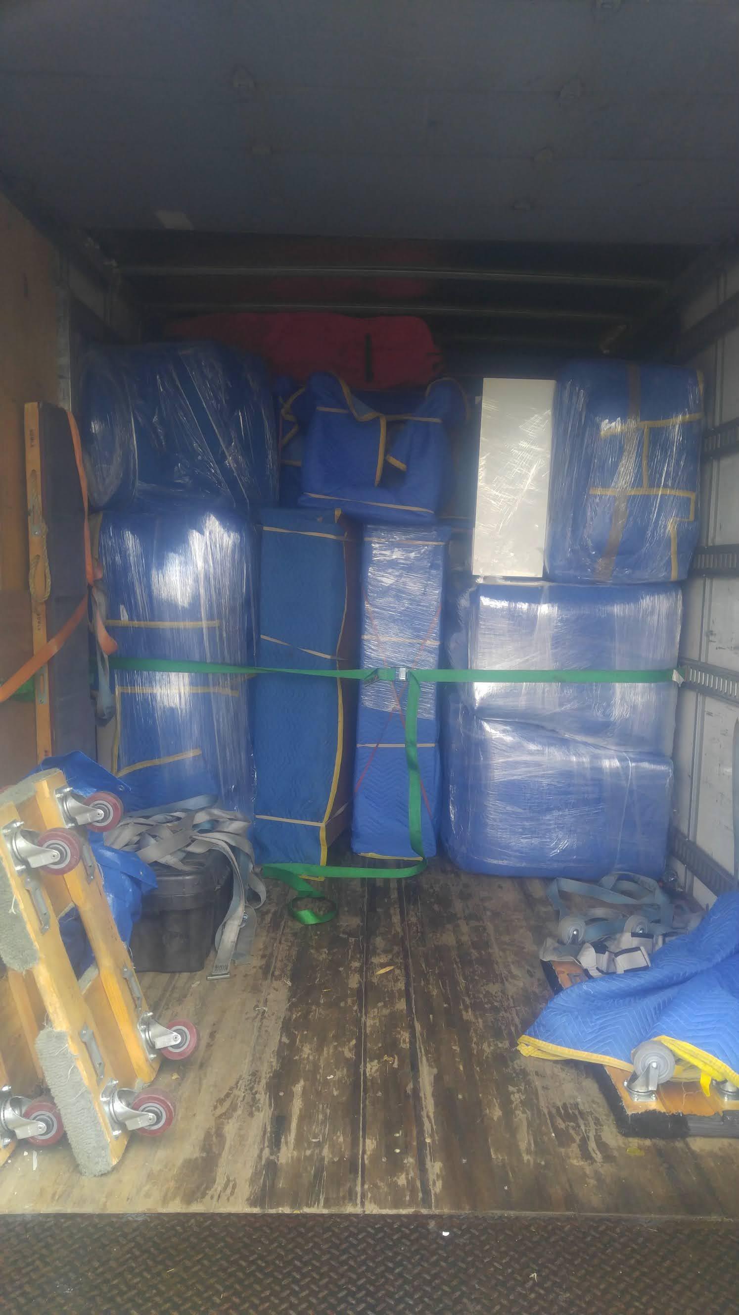 Truck interior with packed furniture, secured with straps and wrapped in blue plastic.