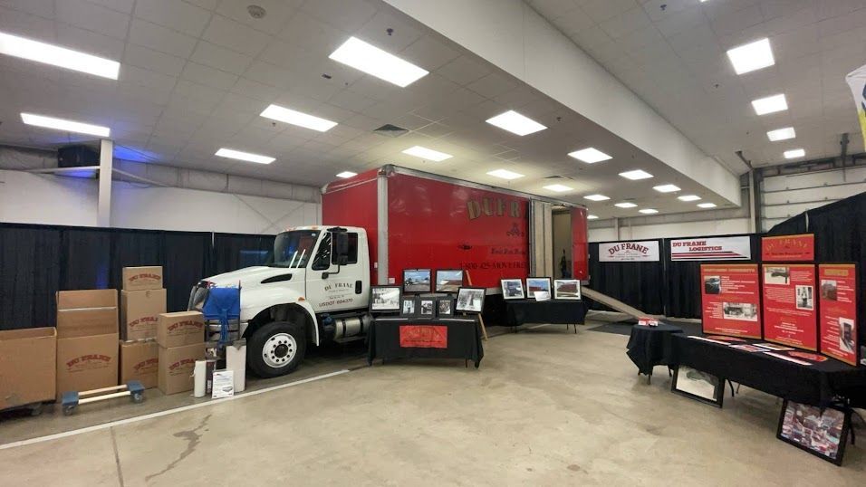 White truck parked in a warehouse beside stacked boxes and red cargo containers on display tables