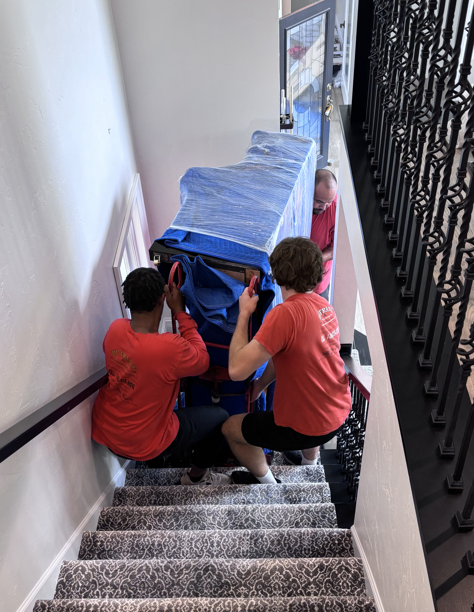 Movers carrying a wrapped piano down a carpeted staircase. Blue padding, red shirts, and white walls.