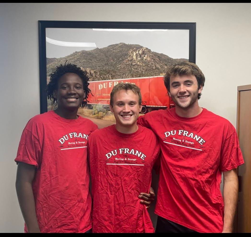 Three people in red t-shirts pose smiling in front of a mountain print.