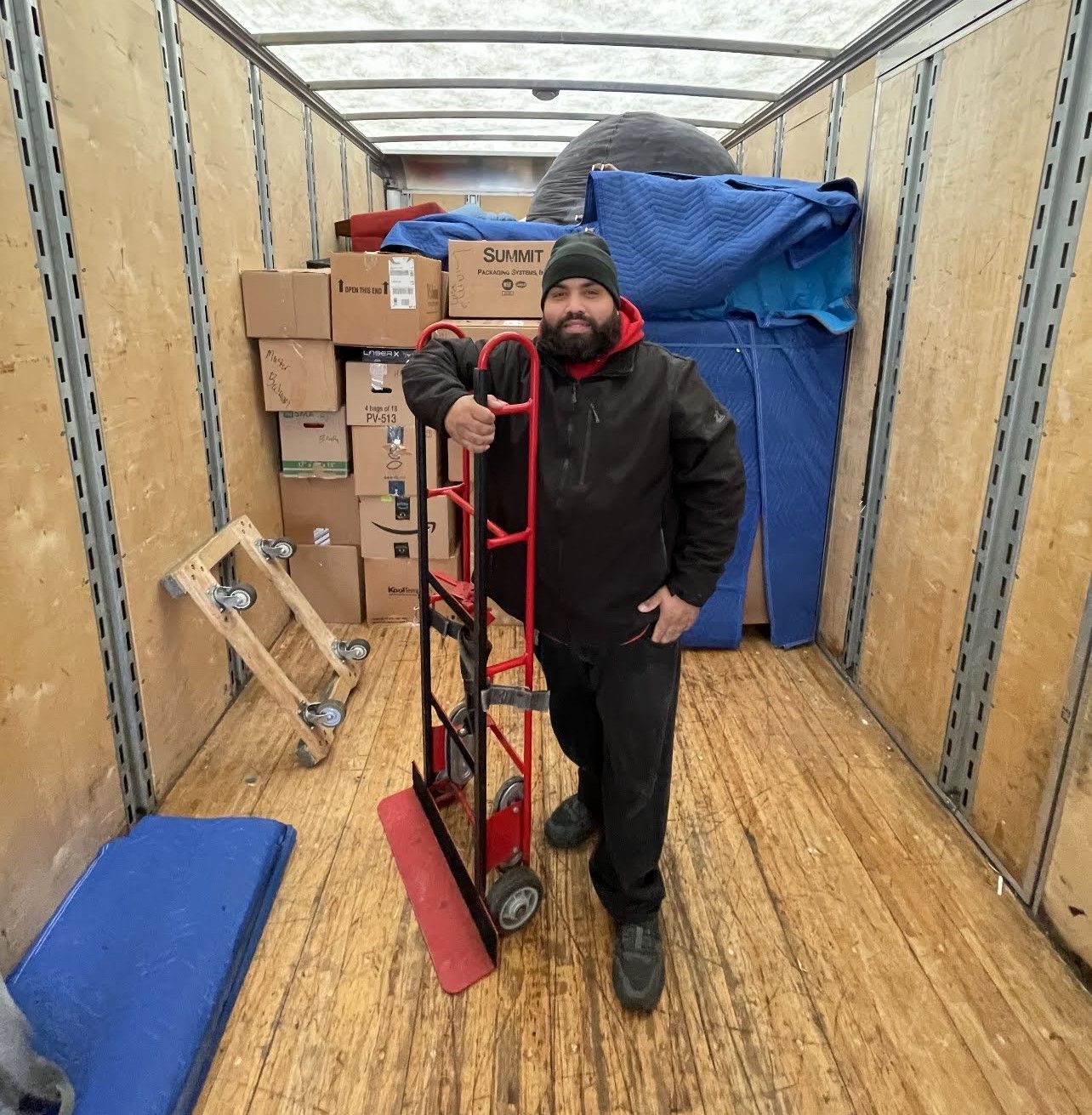 Man in a truck, holding a hand truck. Truck is partially loaded with boxes and furniture pads.