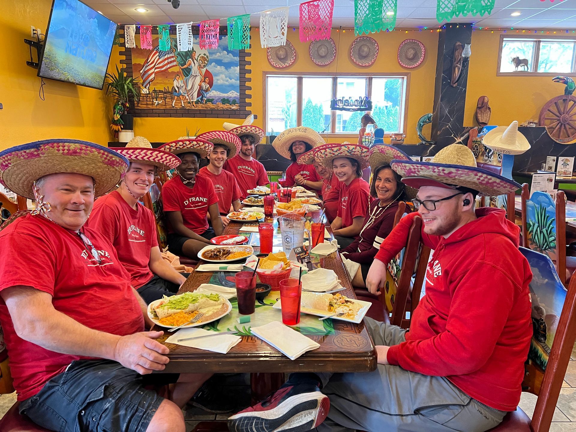 Group of people wearing sombreros and red shirts at a Mexican restaurant, dining together.