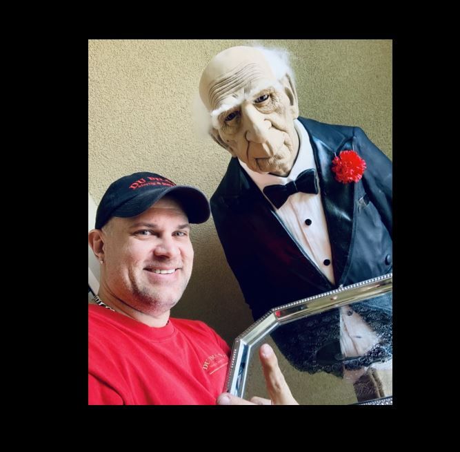 Man in red shirt points at an elderly-looking figure in a tuxedo with a red flower, both indoors.