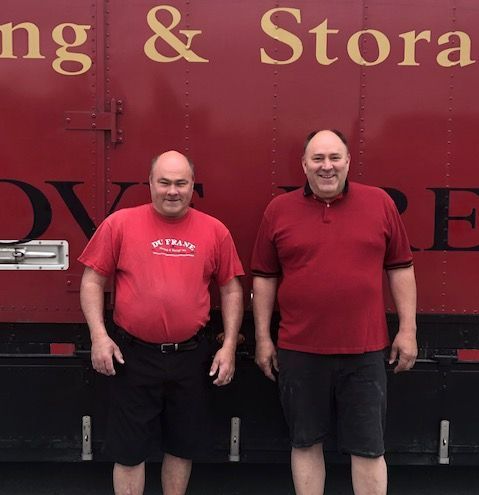 Two men in red shirts and shorts stand in front of a red moving truck with gold lettering.