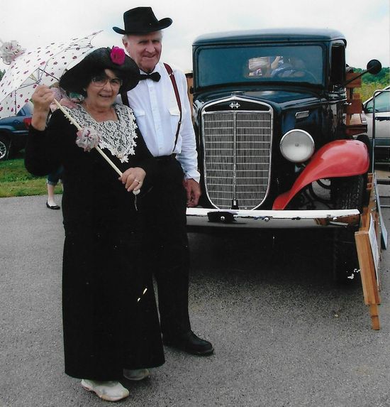 Couple in vintage attire pose by a classic black truck; woman holds parasol.