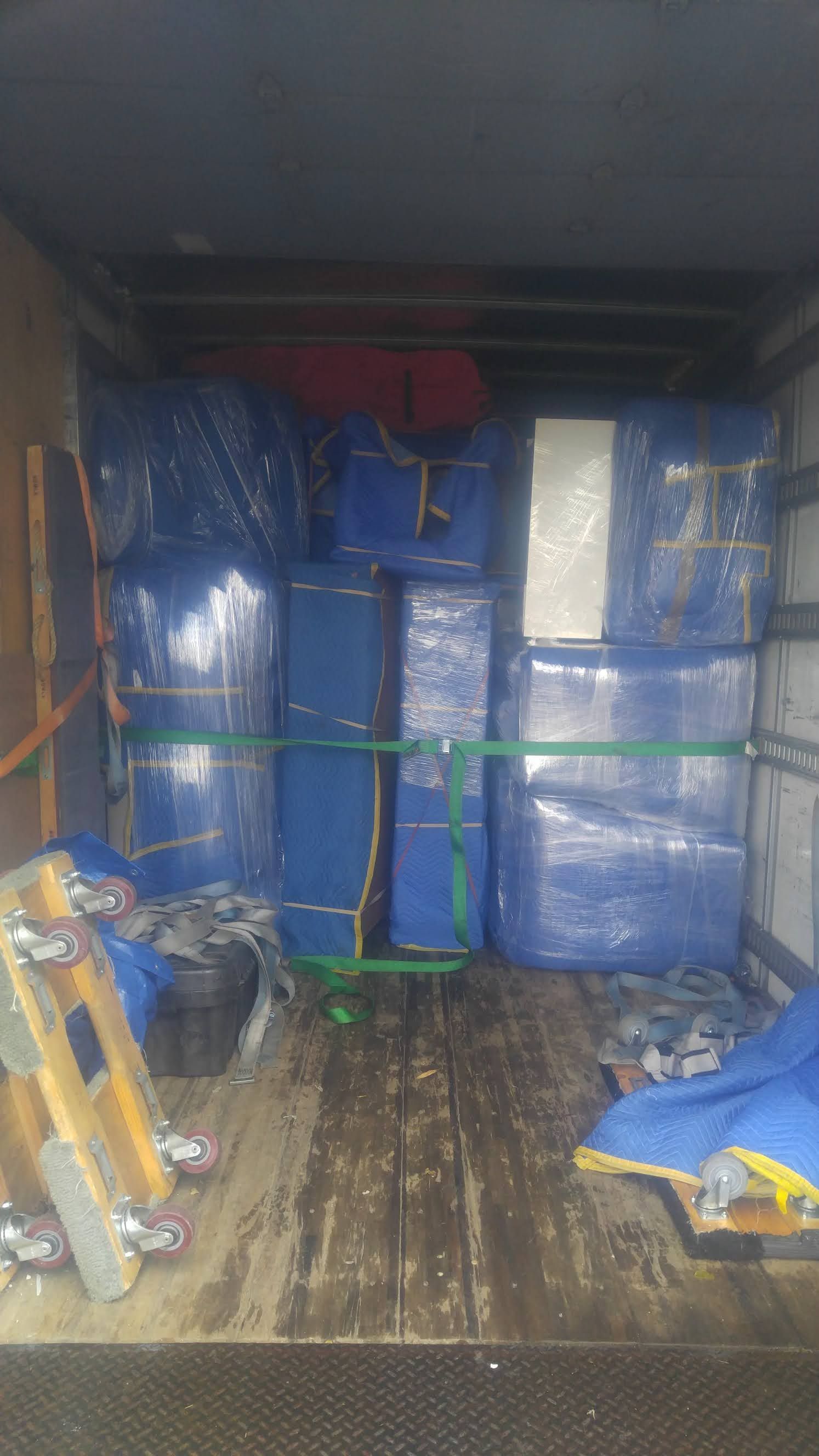 Boxes wrapped in blue plastic and secured in a truck for moving, with a pallet jack visible.