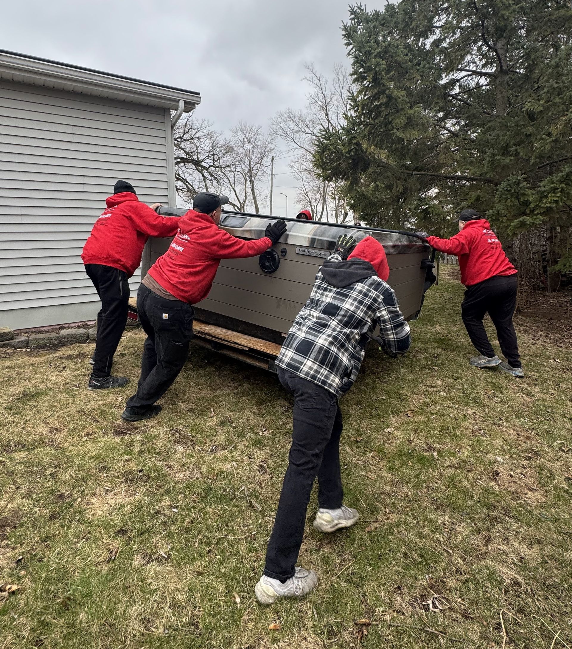 Four people in red jackets and a person in a plaid jacket lift a large, dark object on a grassy area near a building.