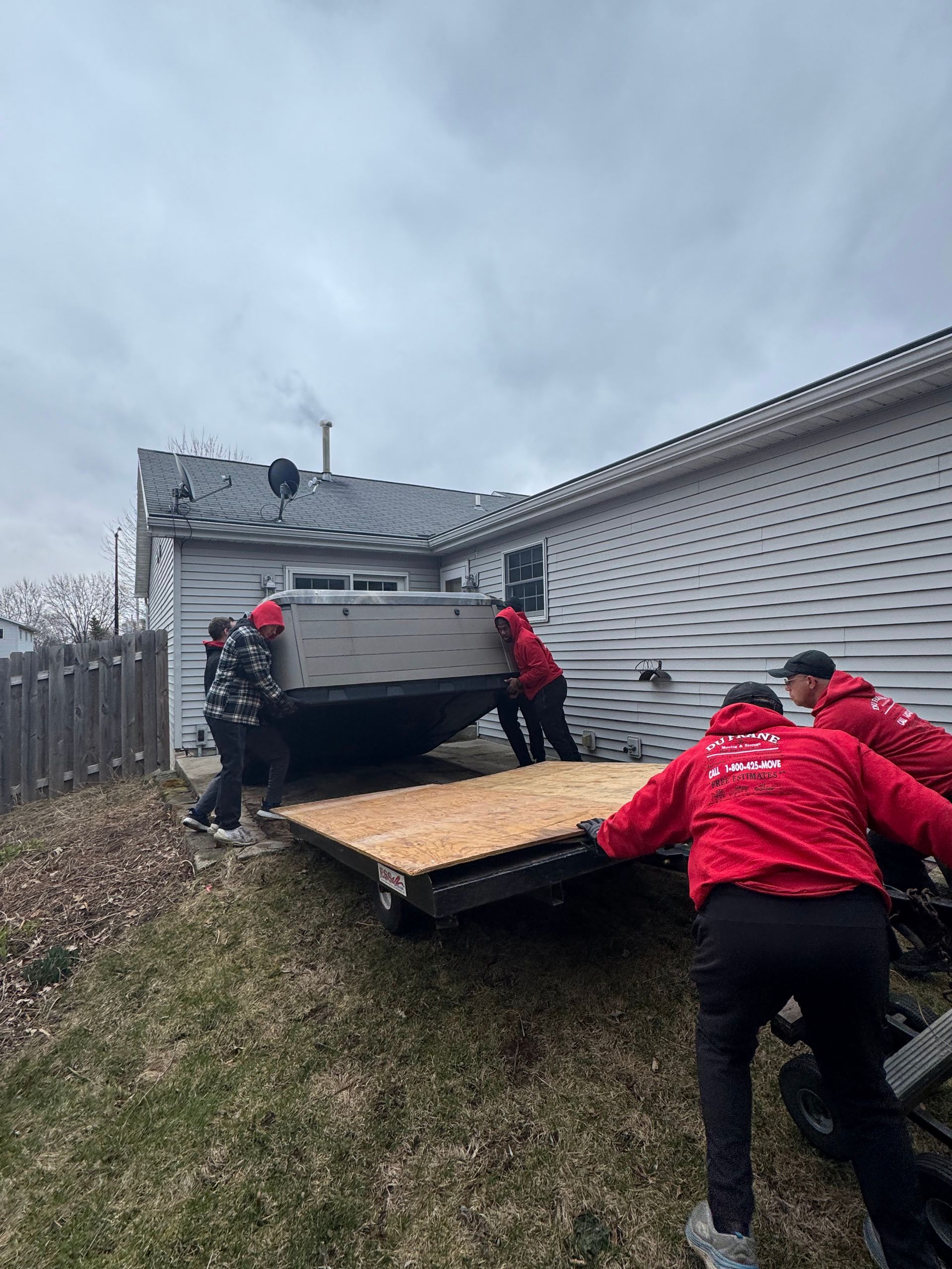 Four people in red jackets lift a large, rectangular object off a trailer next to a house.