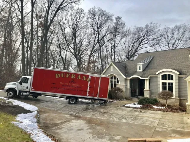 Red moving truck parked in the driveway of a light gray house; bare trees in the background, a bit of snow on the ground.